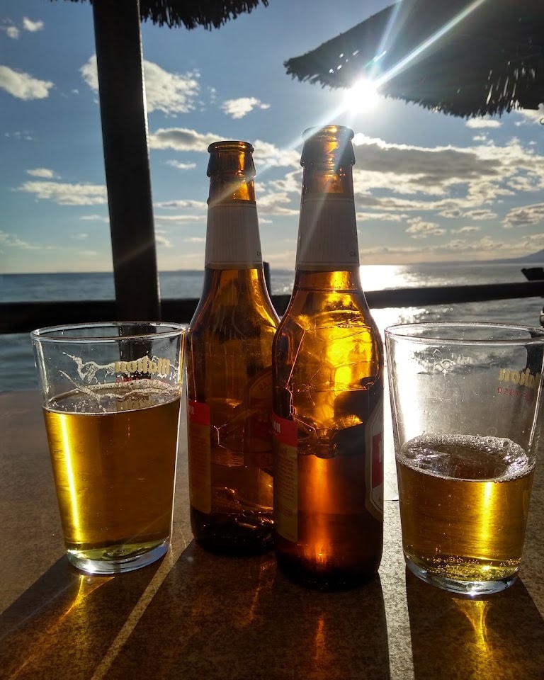 Dos botellas de cerveza y vasos llenos capturan los reflejos dorados del atardecer en la playa.